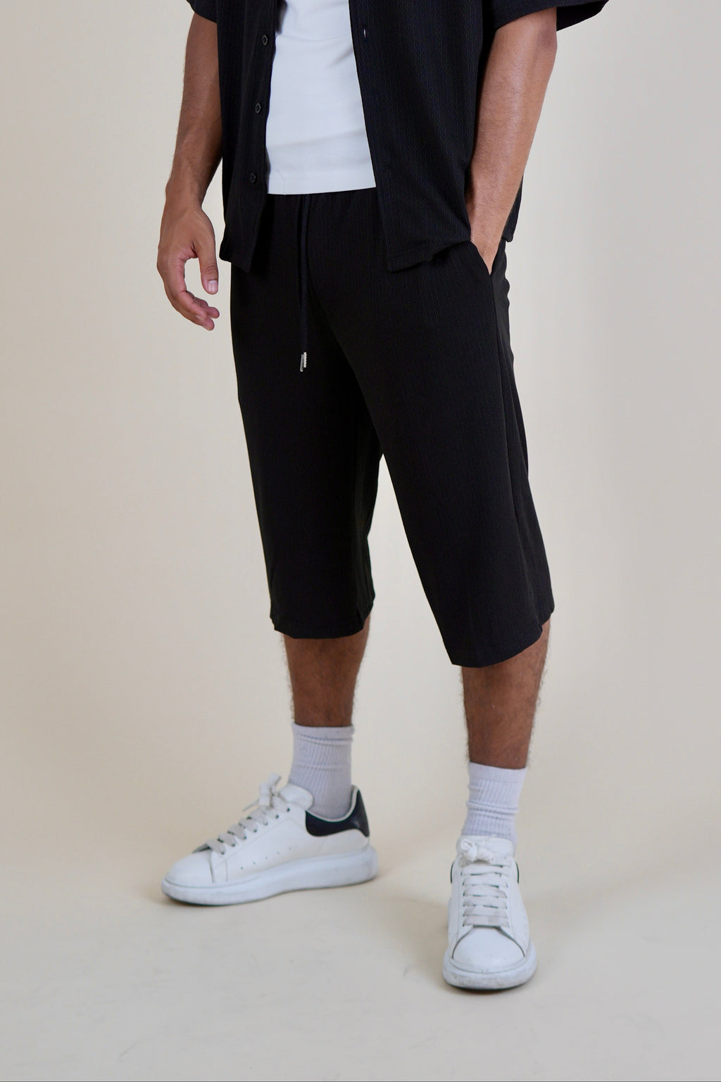 A man wearing a black short-sleeve button-up shirt over a white t-shirt, paired with black drawstring shorts, white socks, and white sneakers, standing against a plain beige studio background.