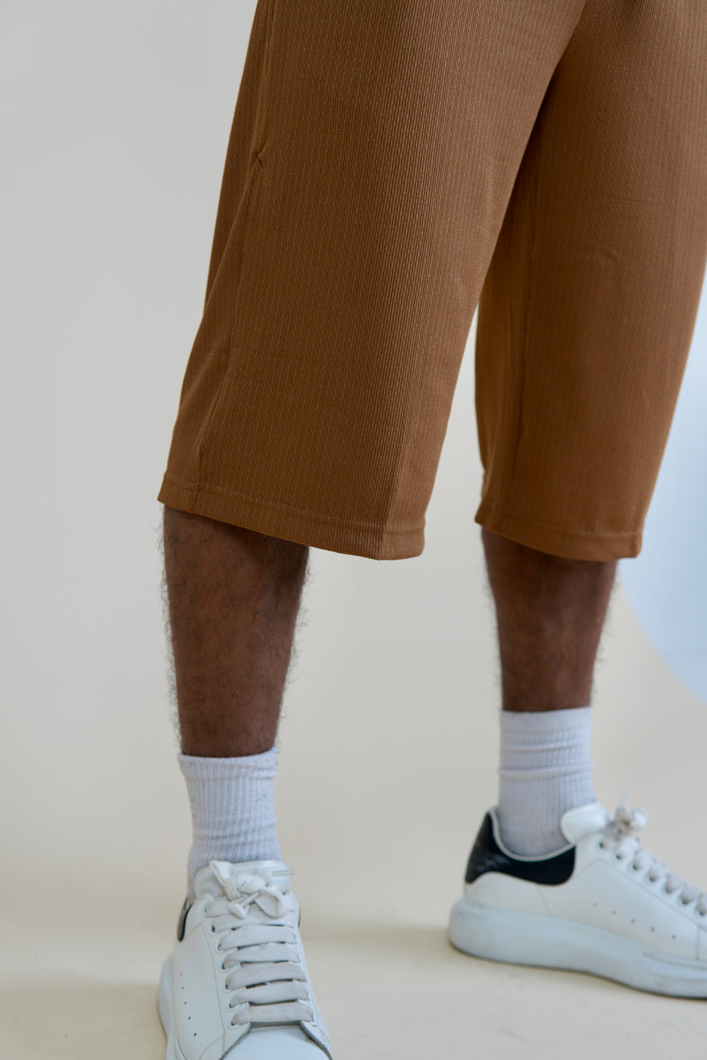 A close-up of a person's legs wearing brown ribbed cropped shorts, white crew socks, and white sneakers with a black heel detail, set against a plain light beige studio background.