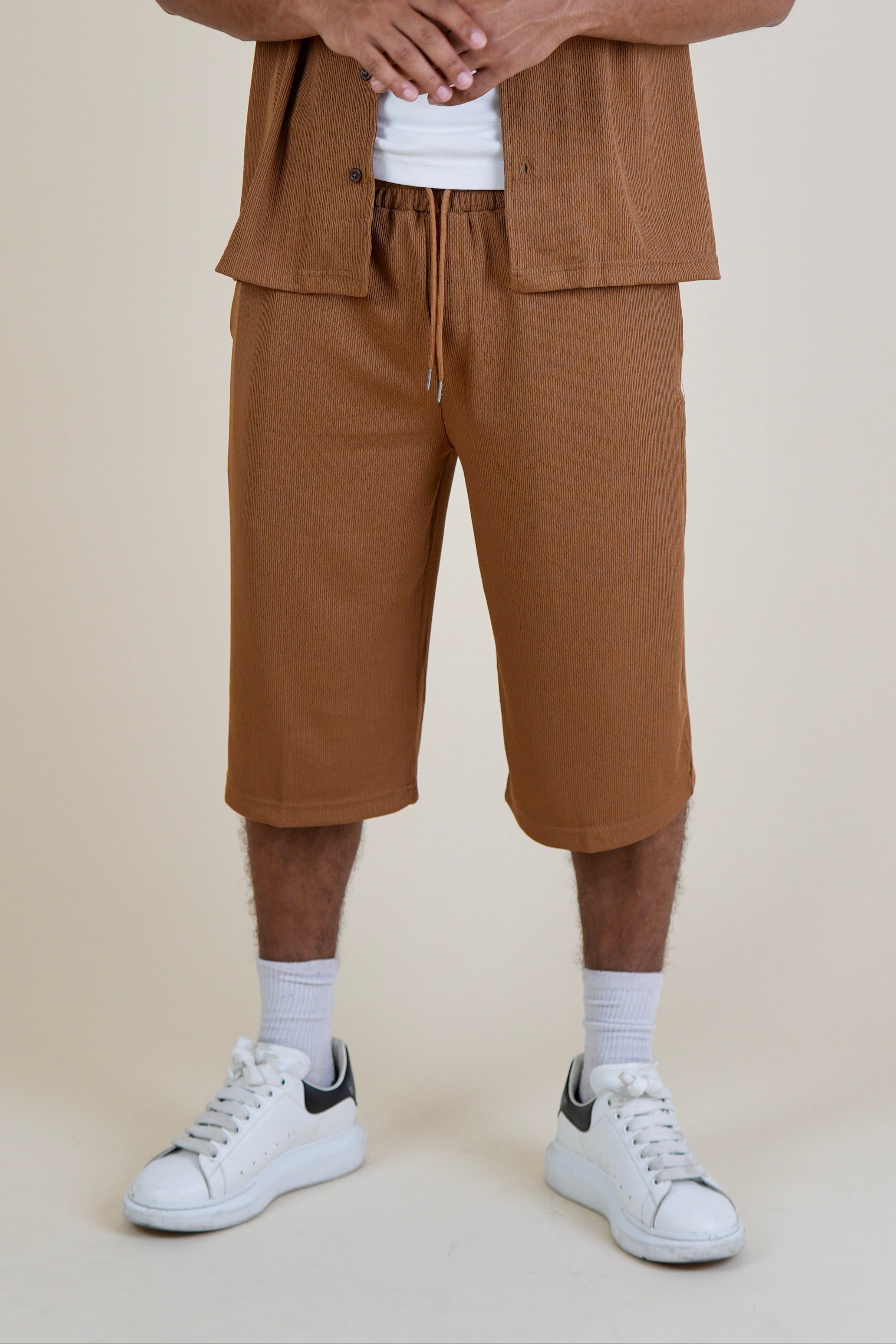 A man modeling a brown short-sleeve button-up shirt and matching brown cropped shorts over a white t-shirt, paired with white socks and white sneakers, against a solid beige background.