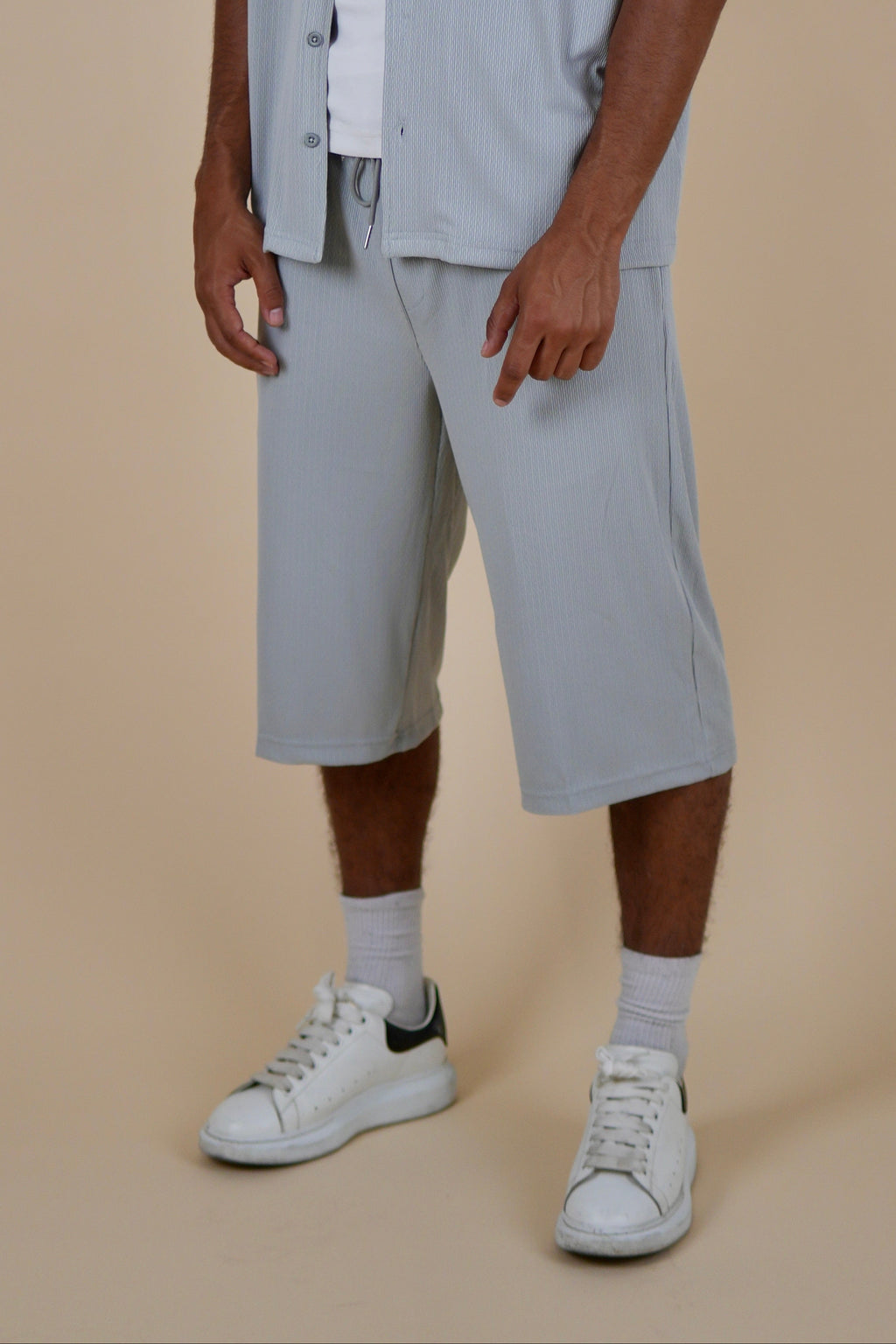A man modeling a light blue-gray short-sleeve button-up shirt, matching cropped shorts, a white t-shirt, white socks, and white sneakers against a solid beige studio background.