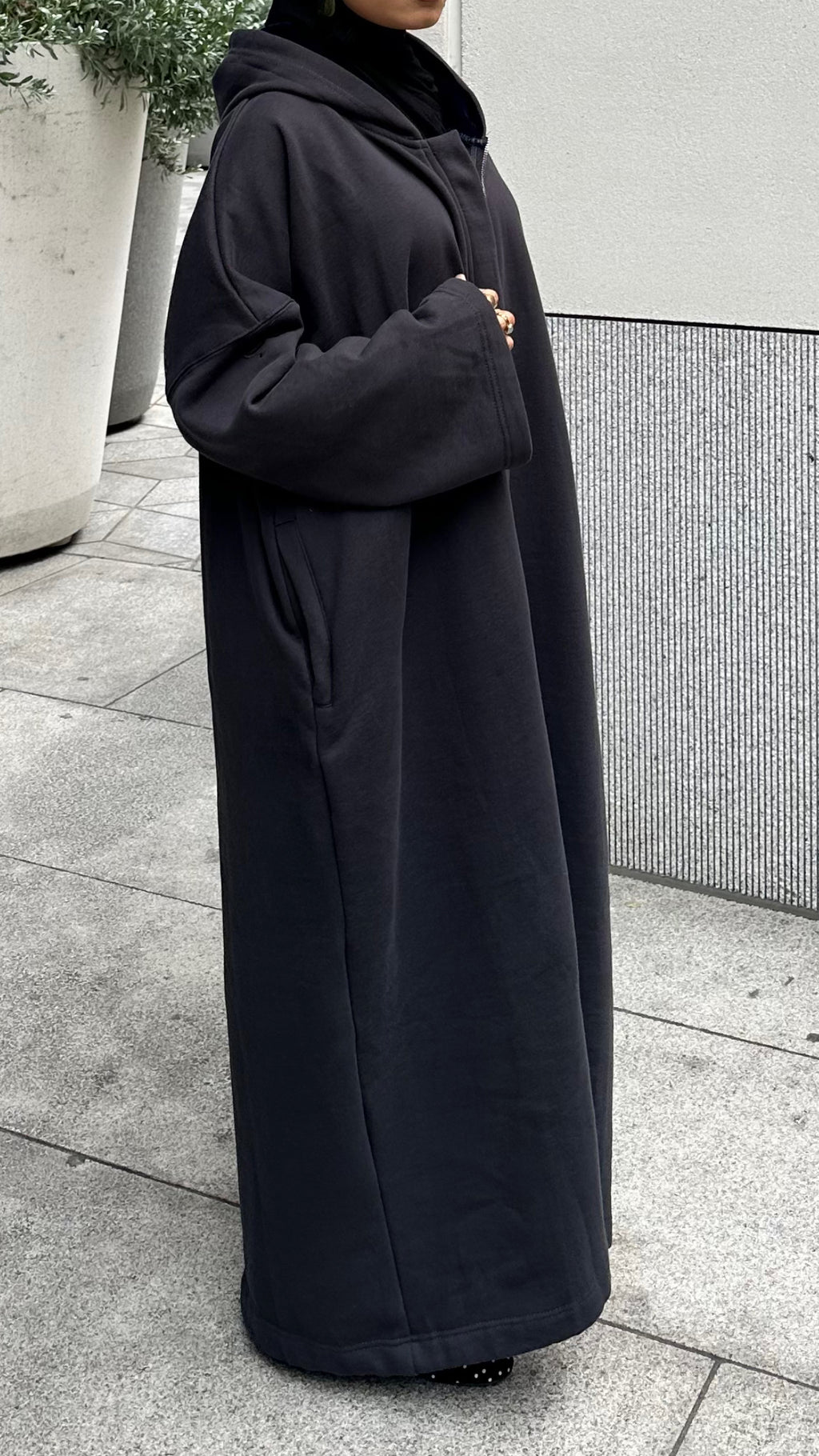 A person wearing a long black hooded coat or abaya standing outdoors on a paved walkway next to a modern wall and planters. Winter abaya perfect for cold winters