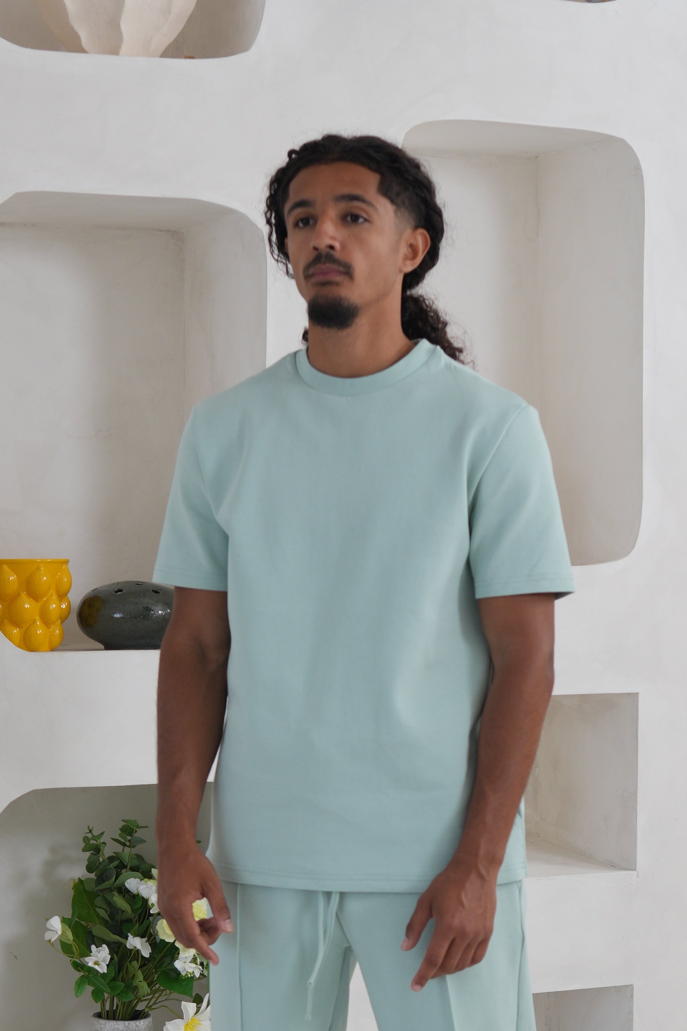 Man wearing a light green outfit standing in a modern interior setting with white walls and minimal decor.