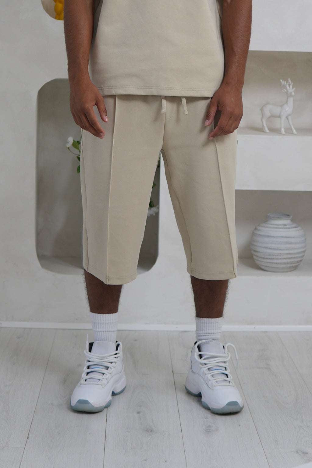 Man wearing a beige outfit standing in a modern, minimalistic room with white walls and light-colored flooring.
