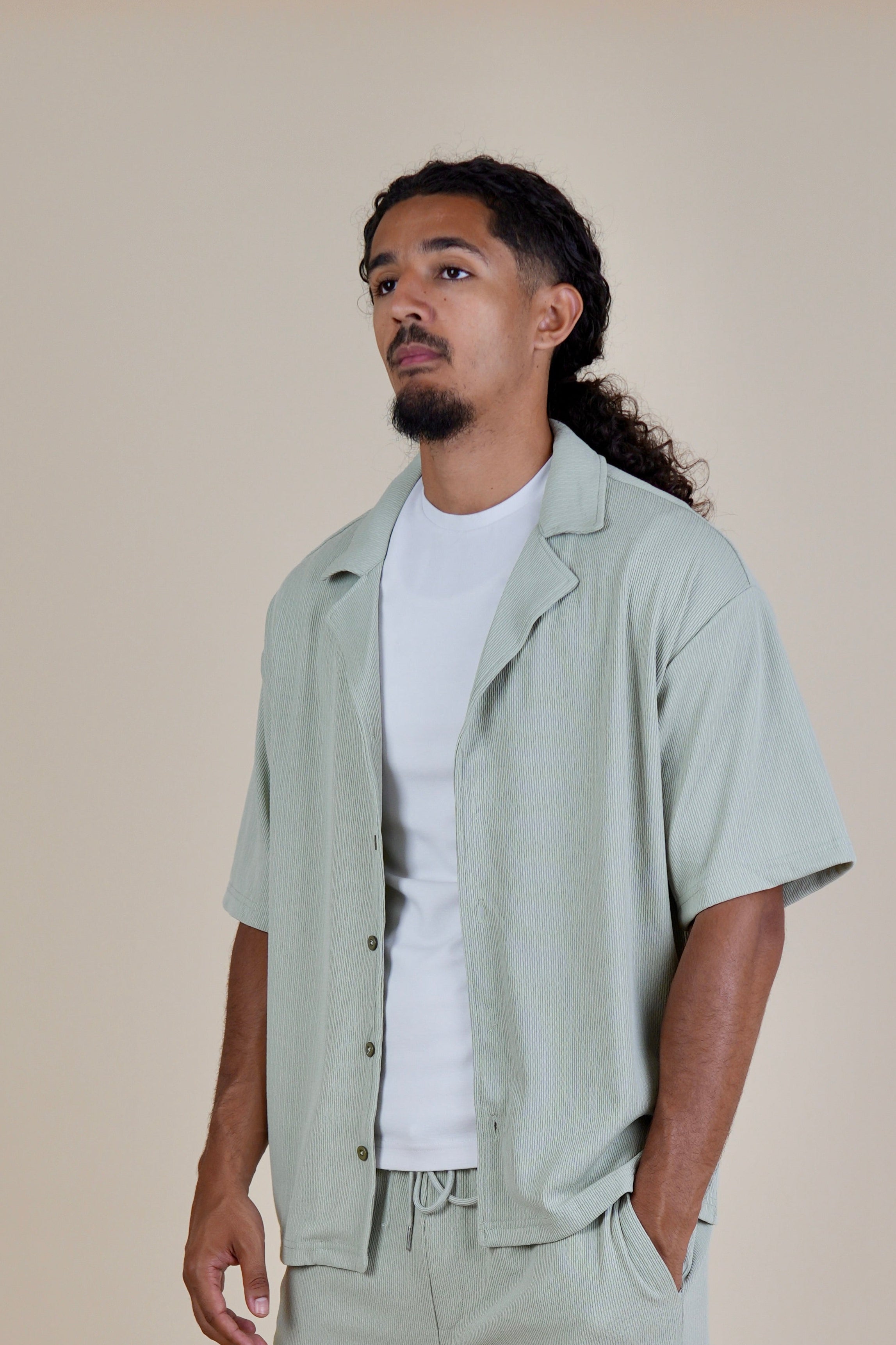 A man modeling a light sage green short-sleeve button-up shirt and matching shorts over a white t-shirt, styled with white sneakers and socks against a solid beige studio background.