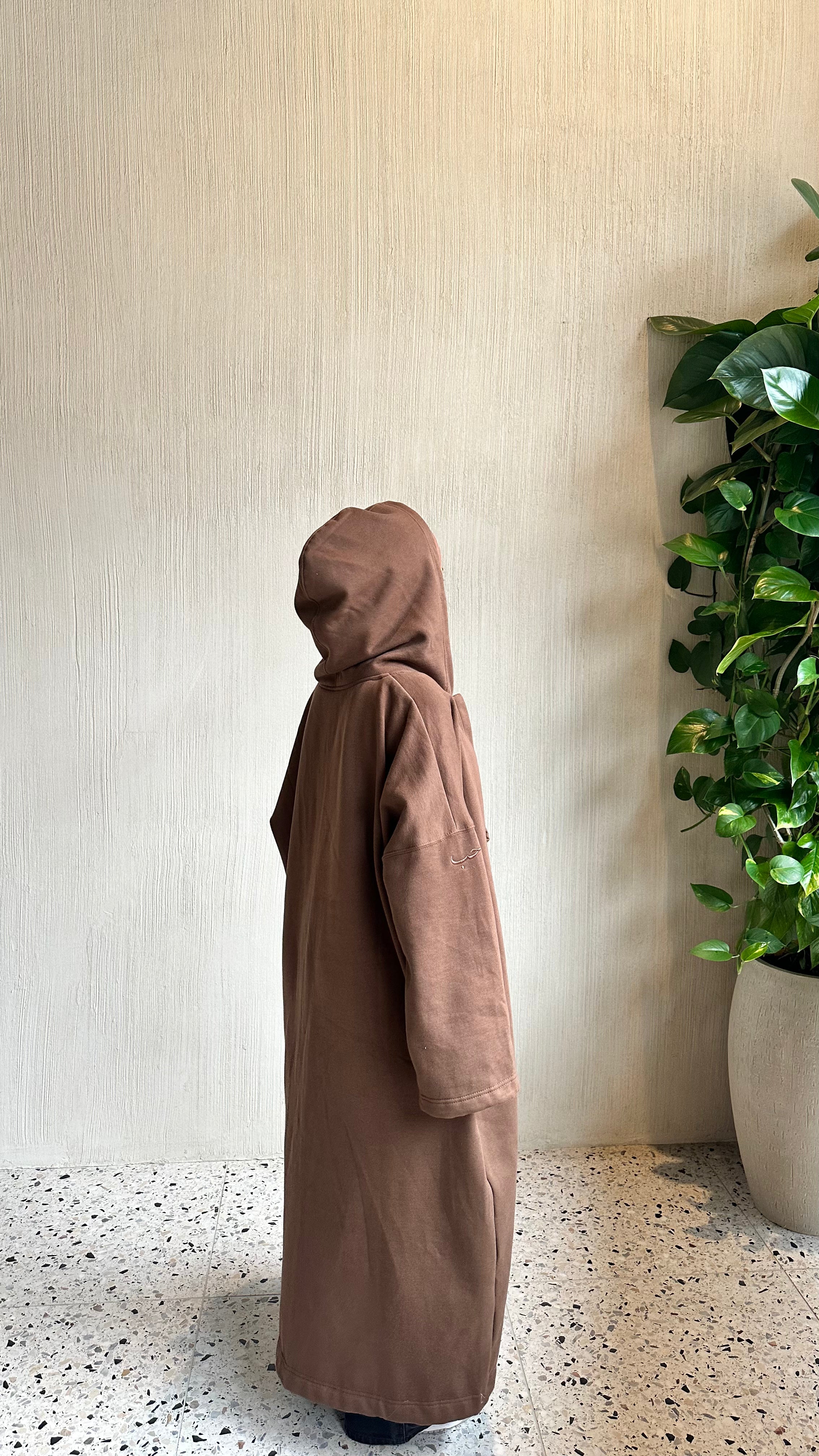 A person wearing a long, loose-fitting brown hooded abaya robe stands in profile against a textured beige wall, with a green potted plant on the right and a speckled floor below.