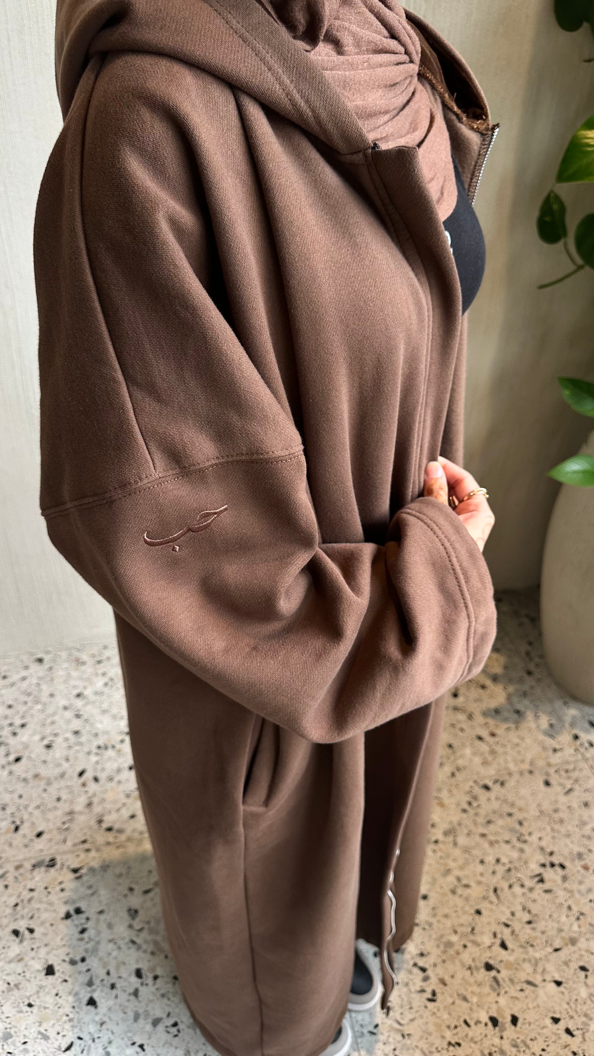 A person wearing an oversized brown hooded sweatshirt or coat, standing indoors on a speckled floor with a plant in the background.