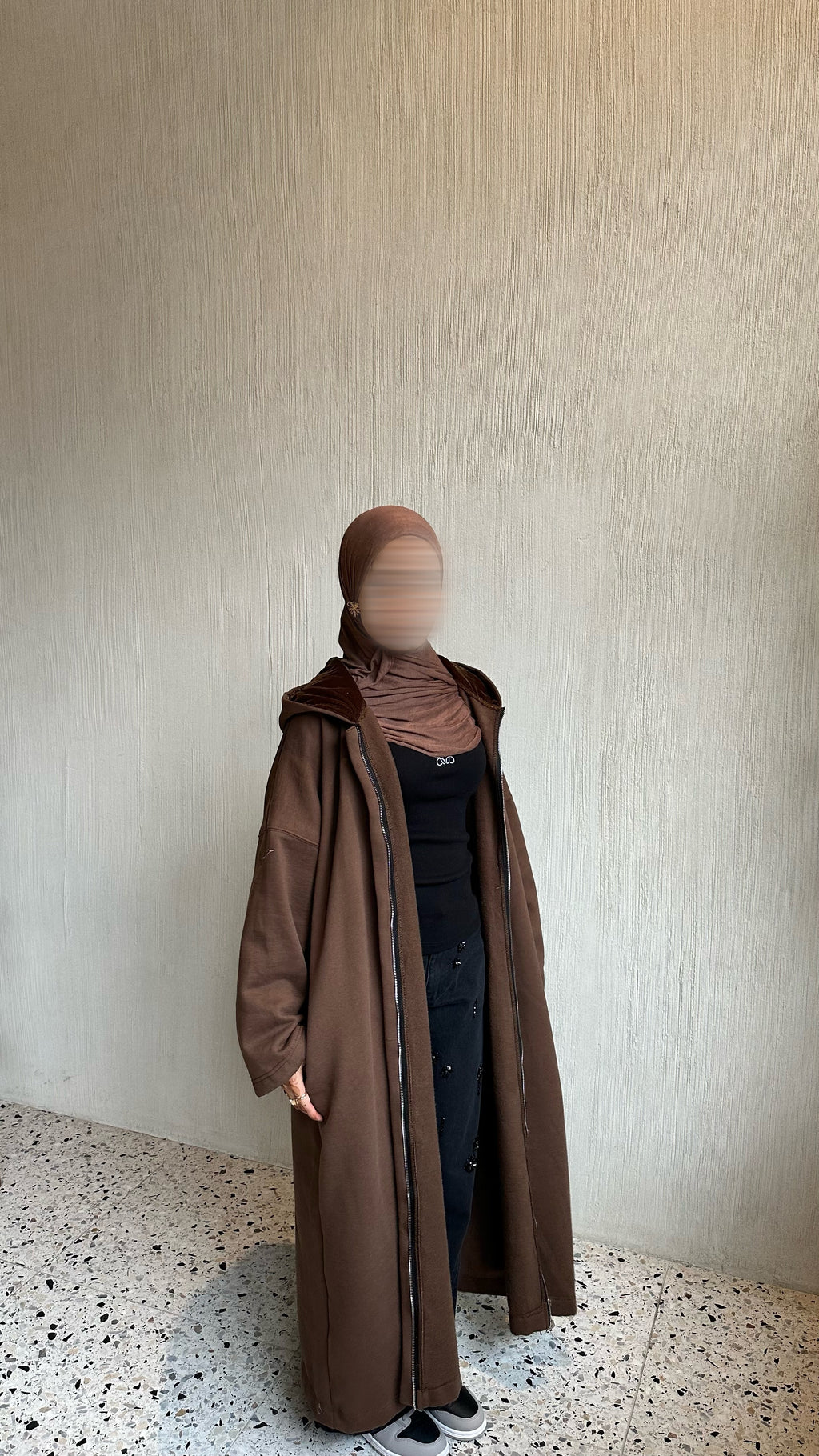A person wearing a long brown hooded coat over a black outfit and a brown hijab, standing against a textured beige wall with a speckled floor. Winter abaya