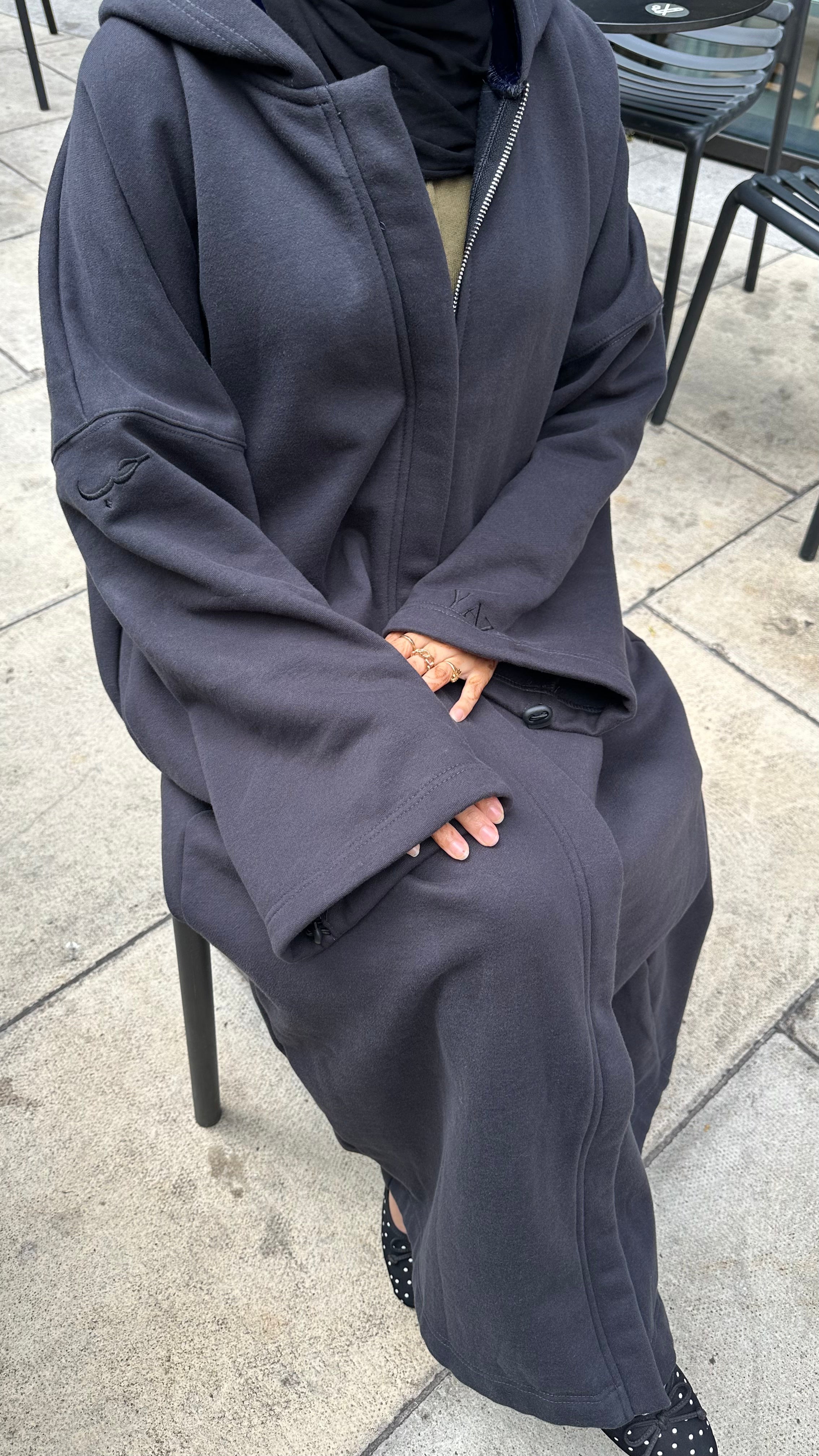 A person seated outdoors wearing an oversized dark charcoal hooded jacket or abaya-style outer layer over a beige top, with a black hijab and black polka-dot shoes visible. The setting is a paved patio with chair legs and outdoor furniture in the background.