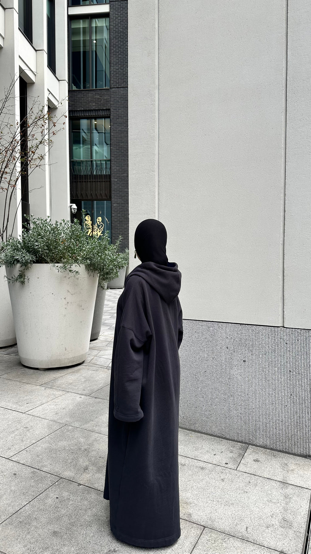 A person wearing a long, loose black hooded robe or coat stands outdoors on a paved walkway beside a modern building wall and large planters. Winter abaya and matching hijab