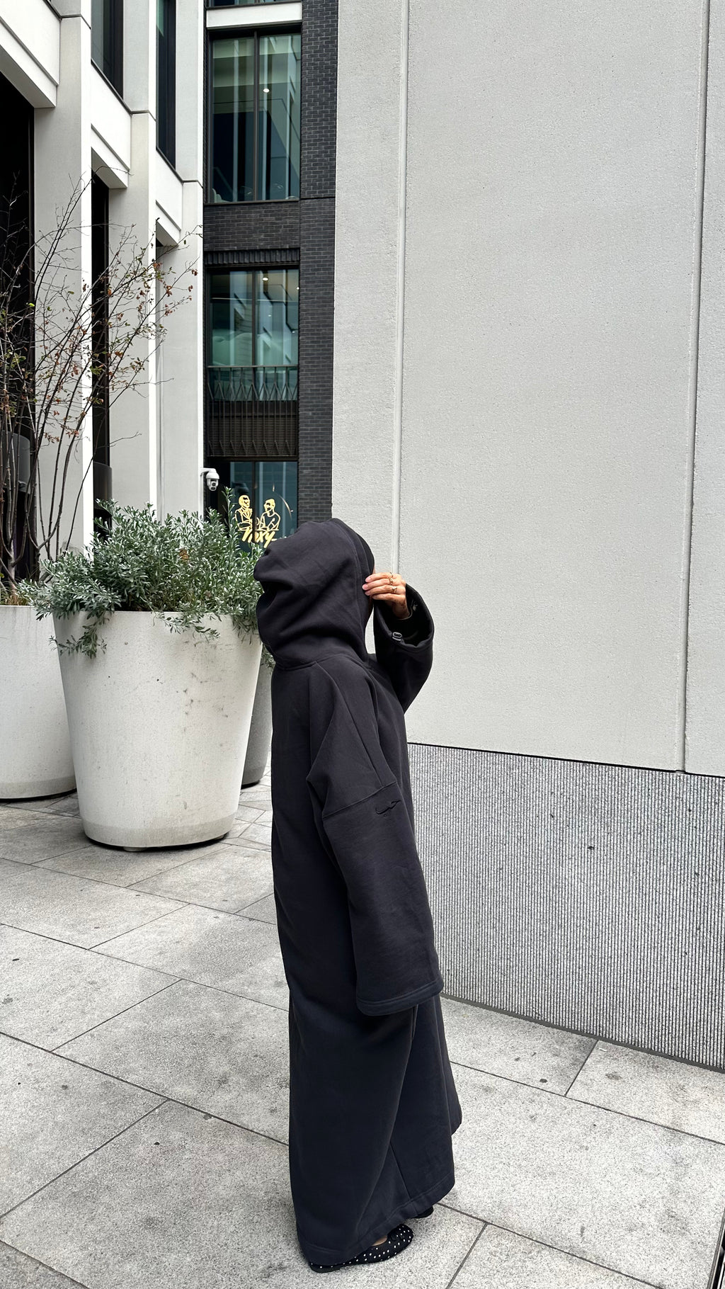 A person wearing an oversized black hooded robe or poncho stands on a paved outdoor walkway beside a modern building with large planters. winter abaya with matching hijab