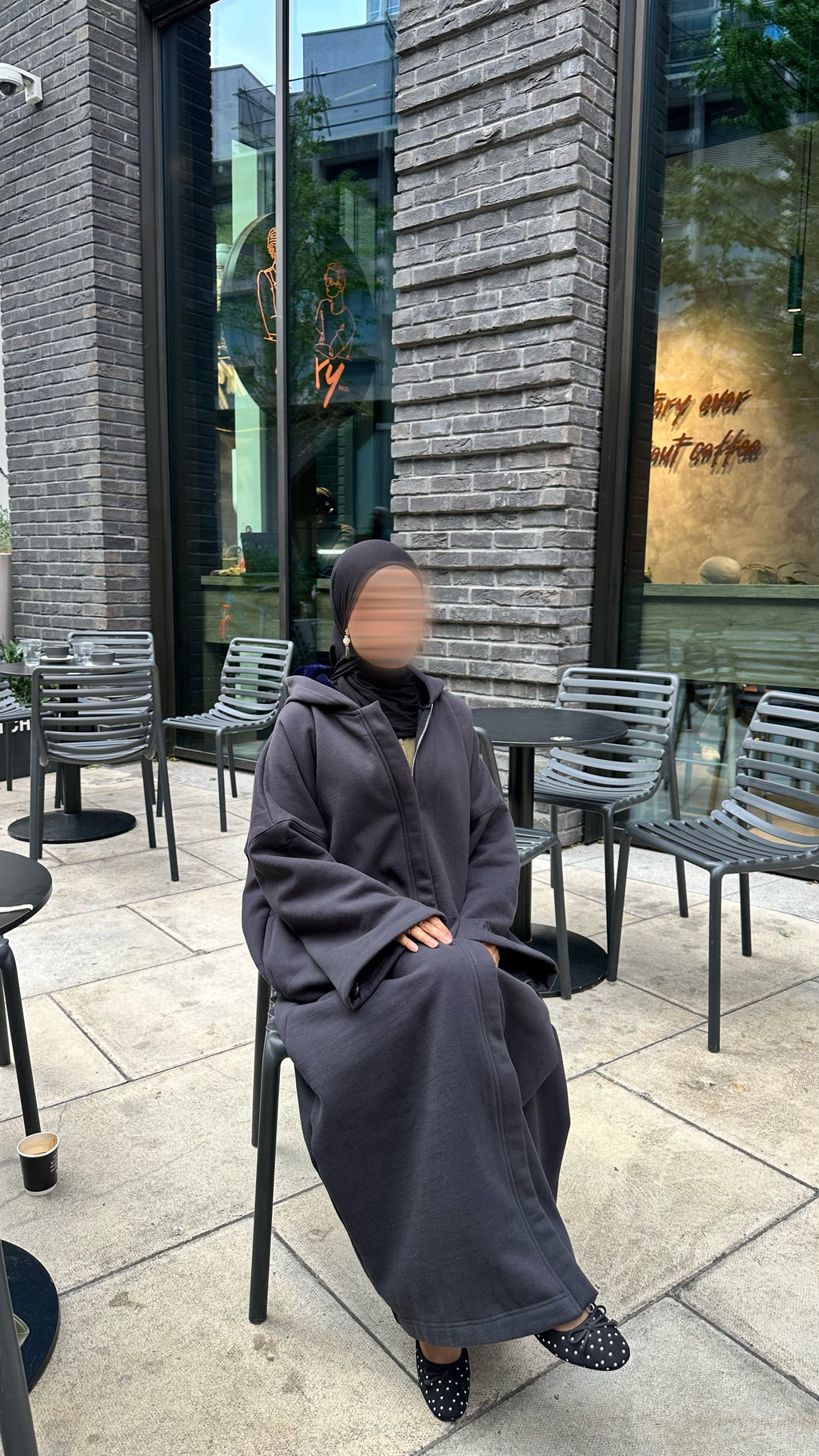 A person wearing a long dark black winter abaya and black hijab sits on a black metal chair at an outdoor cafe table. The background shows a gray brick wall, large glass windows, and other cafe chairs and tables.