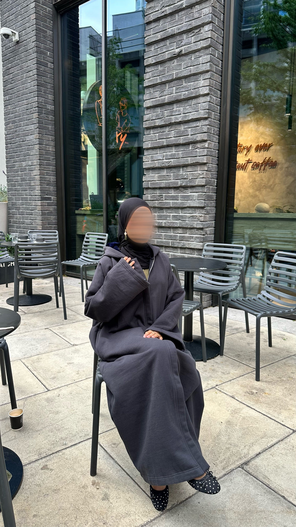 A person wearing a dark gray full-length hooded robe and black hijab sits on a black metal chair at an outdoor cafe table on a stone patio in front of a gray brick building.