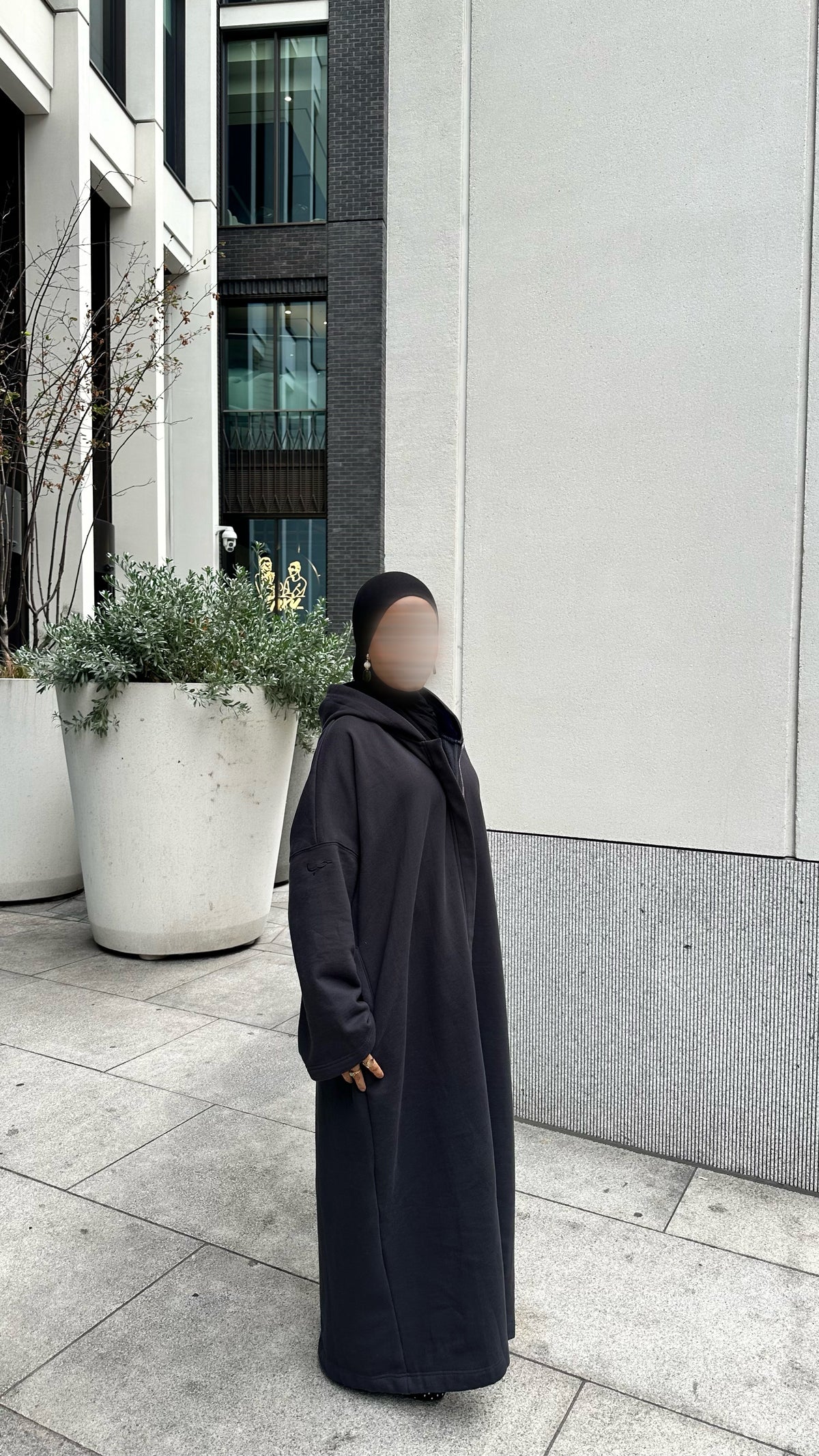 A person wearing a long black hooded coat and black head covering stands outdoors on a paved walkway beside a modern building with large planters. Winter abaya