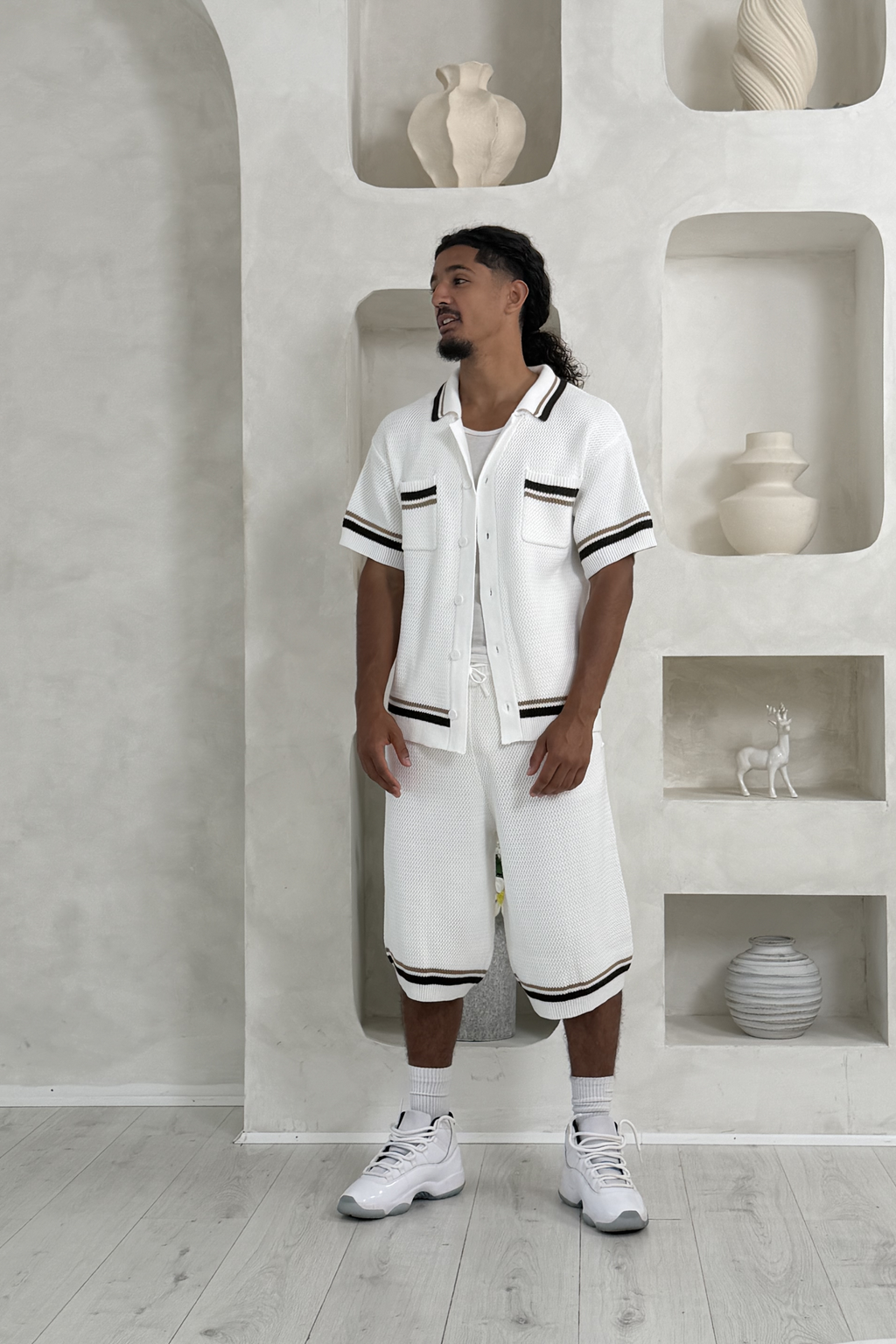Man wearing a white outfit with black accents in a minimalistic room with shelves and decorative items. Knee Length Shorts YAZIR