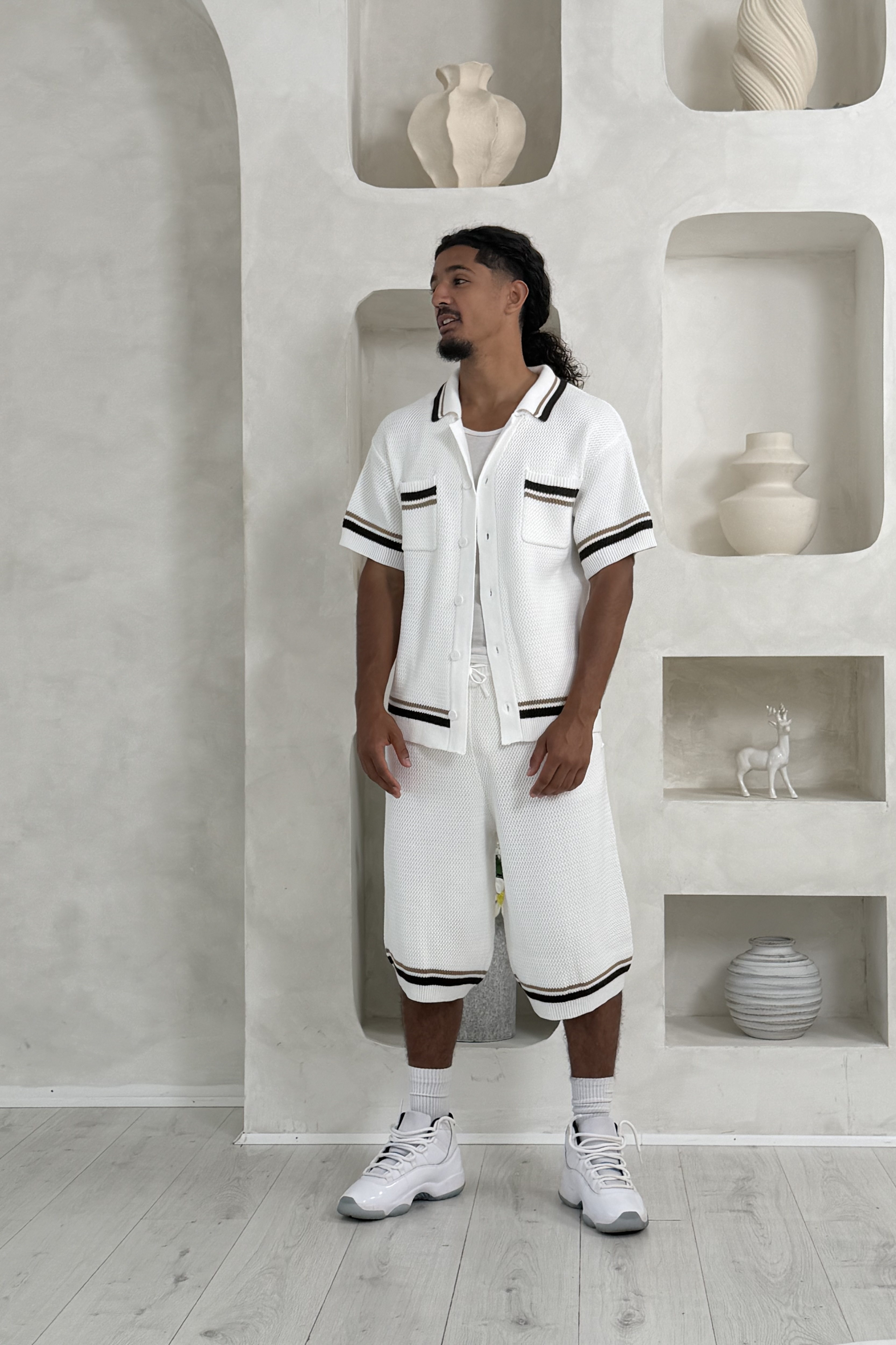 Man wearing a white outfit with black accents in a minimalistic room with shelves and decorative items. Knee Length Shorts YAZIR